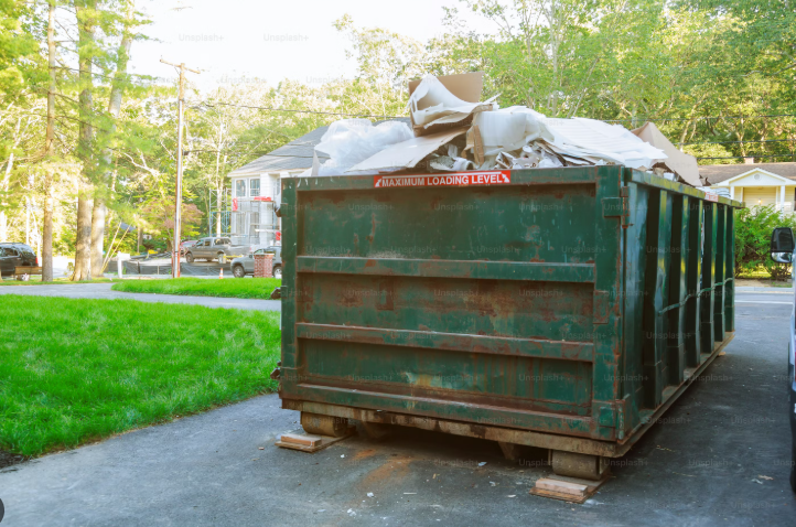 A large rusty dumpster filled with trash bags outside a residential area.